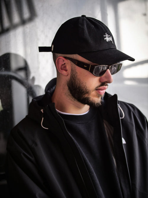 Le rappeur Coelho veste noire et casquette noire, la tête basse, masqué par des lunettes de soleil